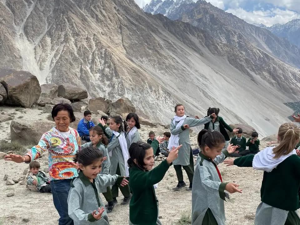 Schoolchildren dancing outdoors on a stony slope with a woman joining in, the valley wall rising behind them