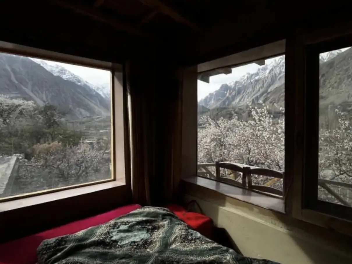 A bed in a corner room, with snow peaks and blossoming trees through the two windows above it